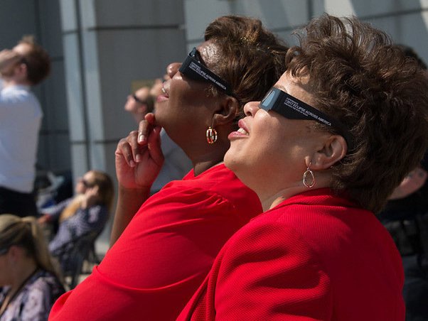 Two people outdoors wearing eclipse glasses and looking up at the Sun during a partial solar eclipse.